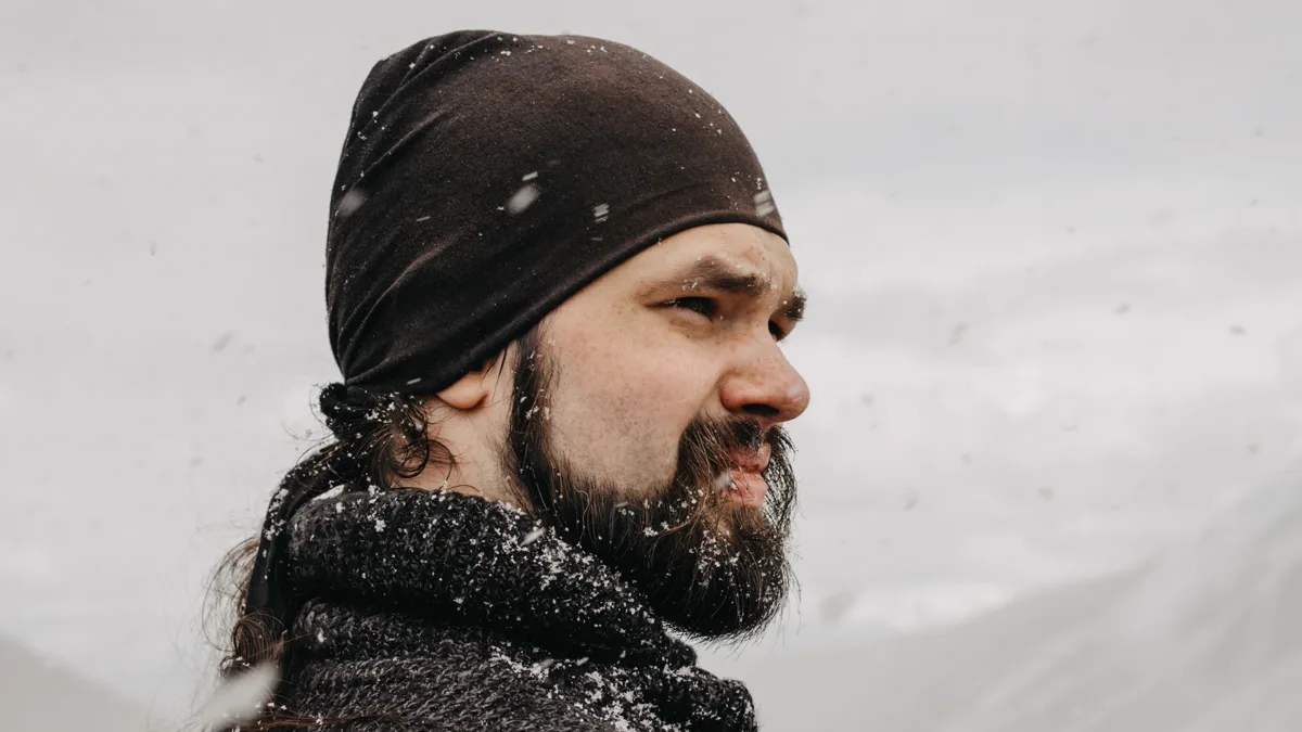 Man with a full beard and long hair wearing a dark beanie and a chunky wool sweater, squinting into falling snow