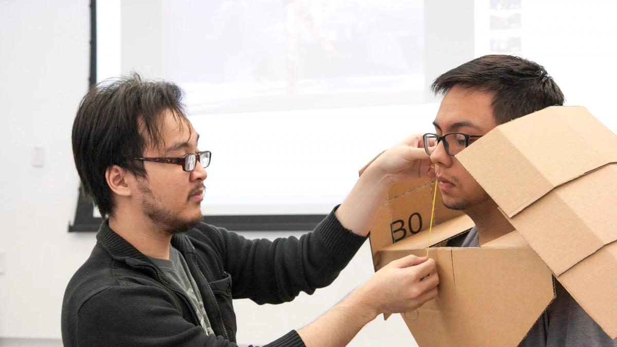 Two students fitting a cardboard chest and shoulder piece cut from a shipping box in a classroom workshop Cardboard armor fitting: one student measures a classmate wearing a chest and shoulder piece folded from a brown shipping box