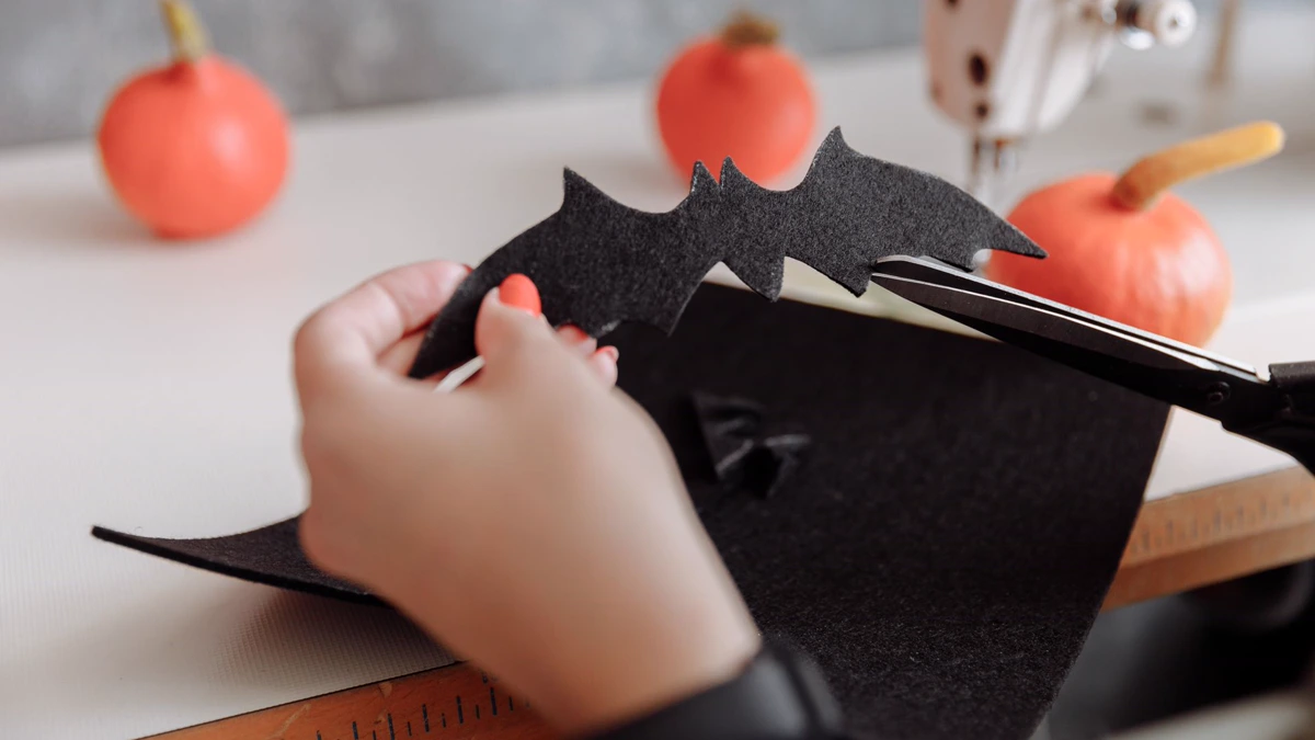 Hands cutting a bat shape from a sheet of black craft felt with scissors, small orange pumpkins on the table behind