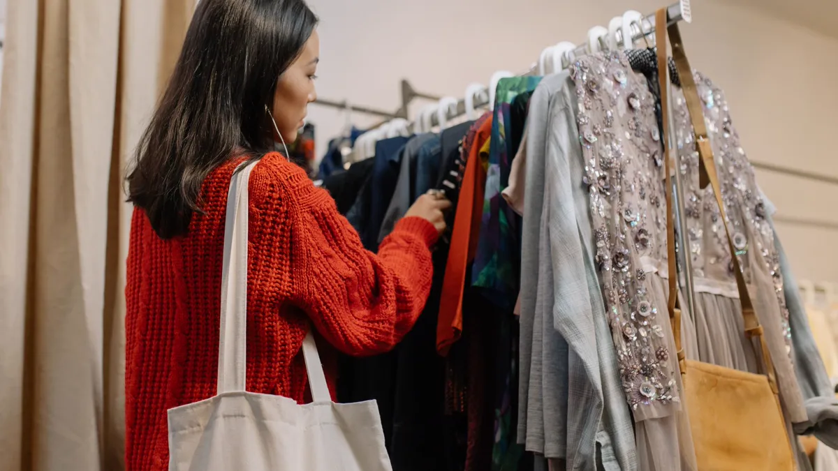 Woman in a red sweater browsing a thrift store clothing rack for cheap Halloween costumes, tote bag on her shoulder