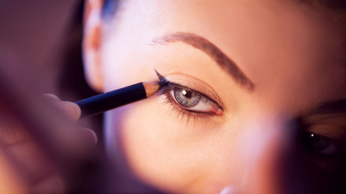 Brown pencil liner drawn along the upper lash line, seen in a mirror Close-up of a brown pencil eyeliner tracing the upper lash line under a softly drawn brow, reflected in a hand mirror