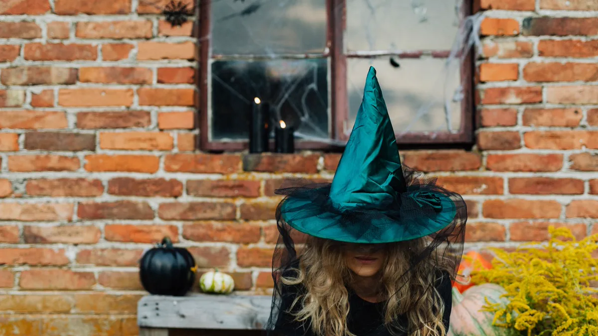 Woman in a plain black top wearing a teal witch hat with a net veil in front of a brick wall with pumpkins