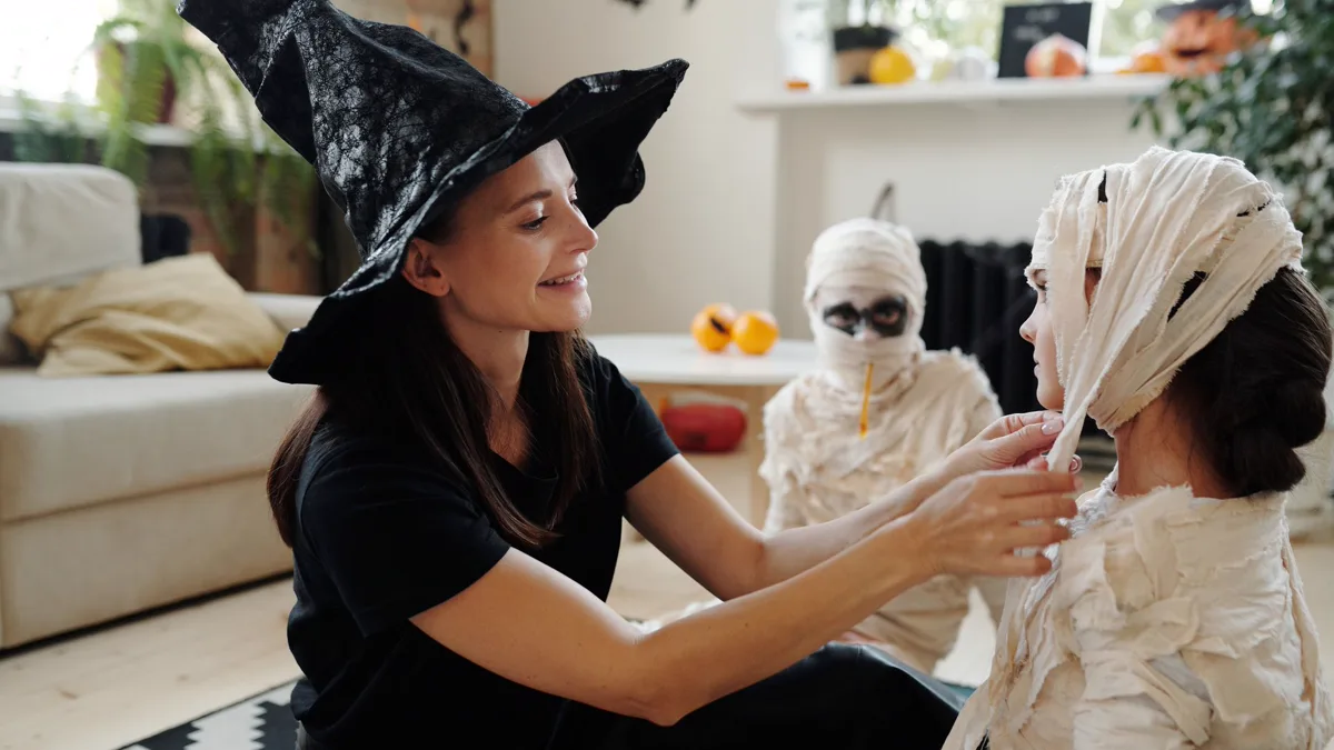 Parent in a witch hat wrapping fabric mummy strips around a child at home Parent in a black witch hat tucking torn fabric strips around a child for a homemade mummy costume, a second wrapped child waiting behind