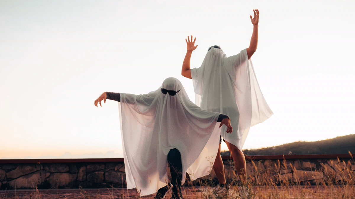 Two people in DIY Halloween costumes made from white bedsheets with sunglasses, posing outdoors at sunset