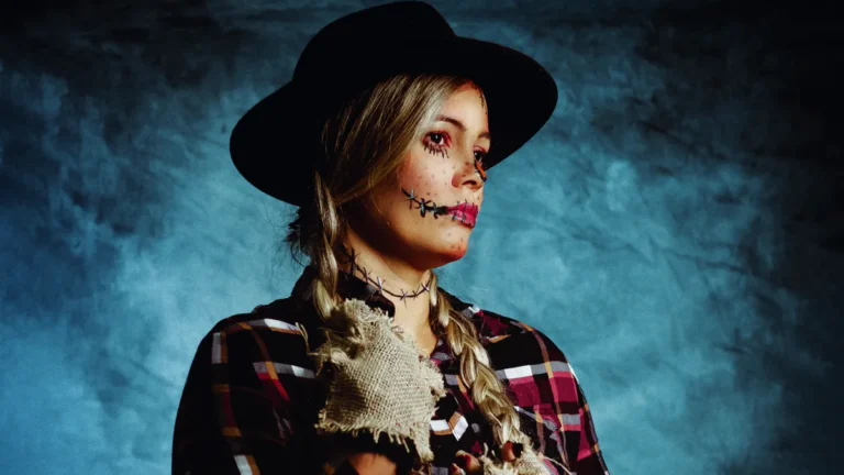 DIY scarecrow costume with a red plaid flannel shirt, a burlap patch at the collar, braids, a black hat and a stitched face
