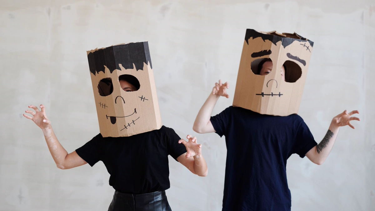 Two people in black T-shirts wearing brown paper bag monster heads with marker faces and a black paper fringe