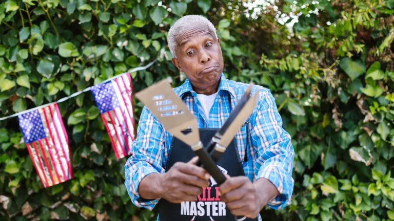 Easy Halloween costumes for men: a man in a blue plaid shirt and black grill apron crossing a spatula and tongs in front of a hedge
