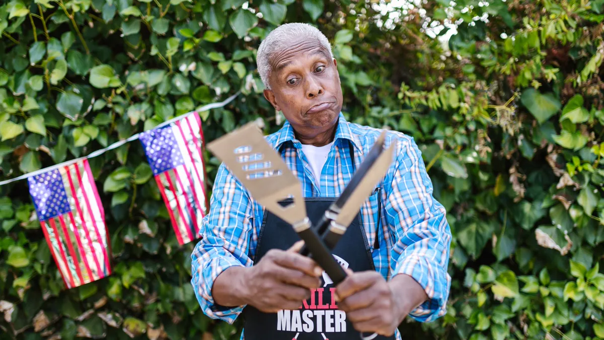 Easy Halloween costumes for men: a man in a blue plaid shirt and black grill apron crossing a spatula and tongs in front of a hedge