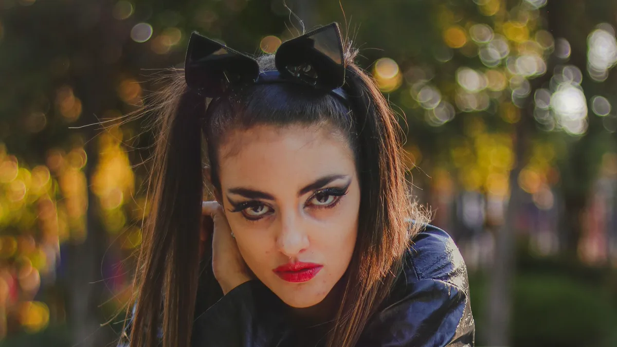 Woman with winged cat eyeliner, red lipstick and black cat ears on a headband, posed outdoors in autumn light