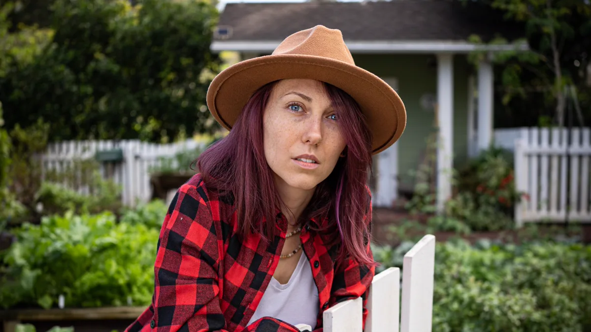 Woman in a red buffalo check flannel shirt and a tan felt wide brim hat leaning on a white garden fence