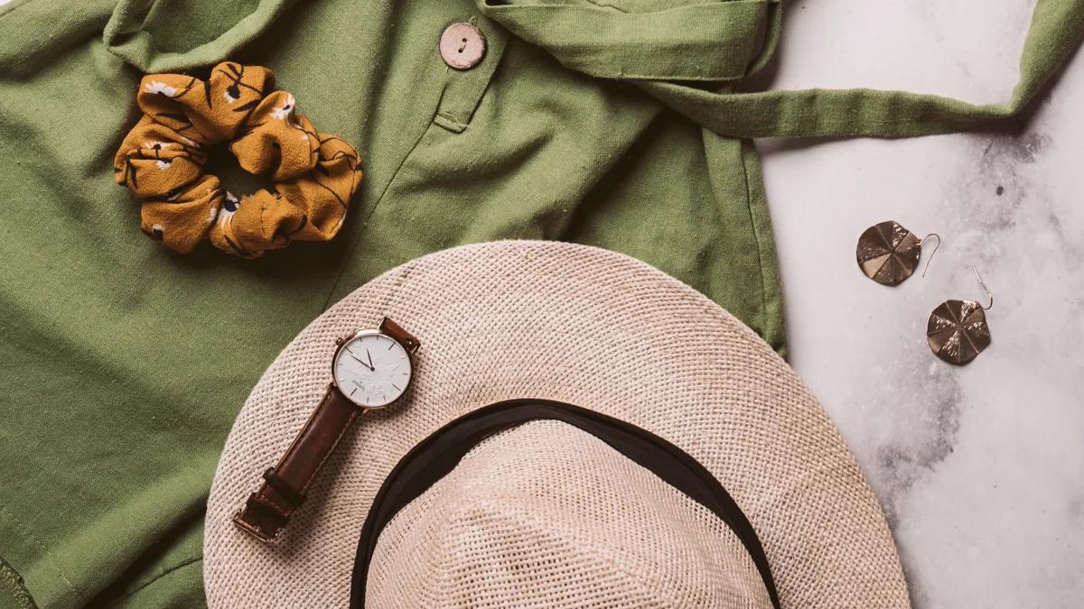 Flat lay of an olive button shirt, straw sun hat, wristwatch and scrunchie Explorer archetype flat lay: olive green button shirt, woven straw sun hat, brown leather wristwatch and a mustard scrunchie on marble