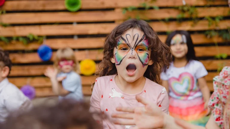 Face paint ideas in action: a curly haired girl with a rainbow butterfly painted across her forehead and cheeks at a backyard party