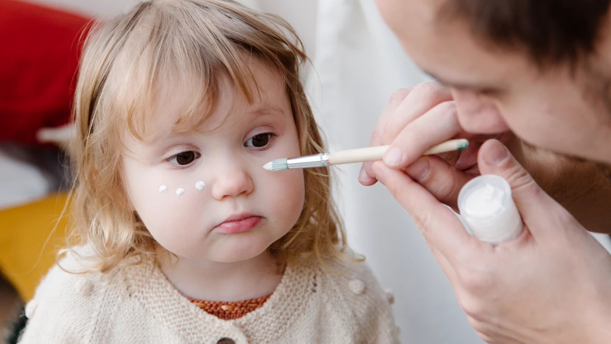 Parent painting white dots on a toddler's cheek with a round brush Parent using a round brush and a small pot of white paint to add a row of dots under a toddler's eye on the cheekbone