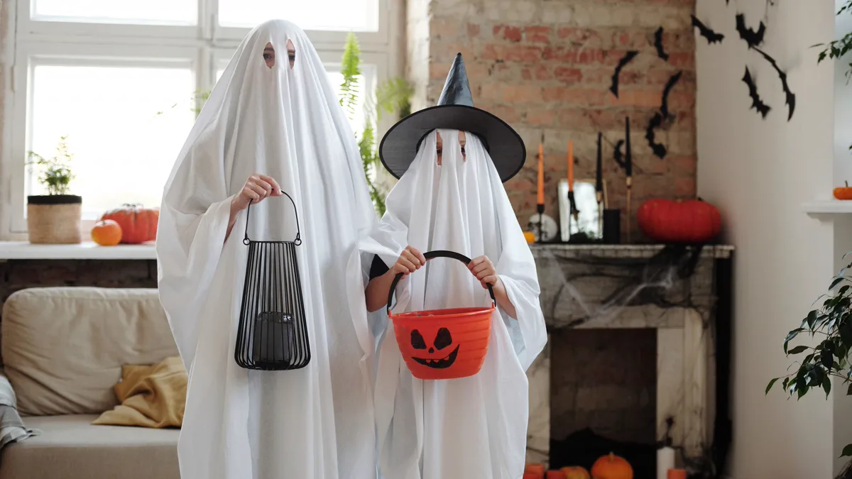 Parent and child in white sheet ghost costumes, the child in a black witch hat, holding a pumpkin bucket Family Halloween costumes made from white sheets: a parent and child dressed as ghosts, the child wearing a witch hat and holding a pumpkin bucket