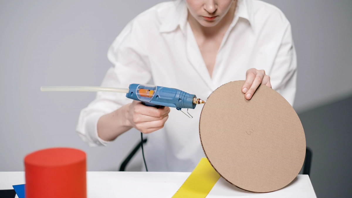 Woman applying hot glue from a blue glue gun to the edge of a round cardboard panel Woman running a bead of hot glue along the edge of a round cardboard disc, the way layers are joined on a DIY cardboard costume