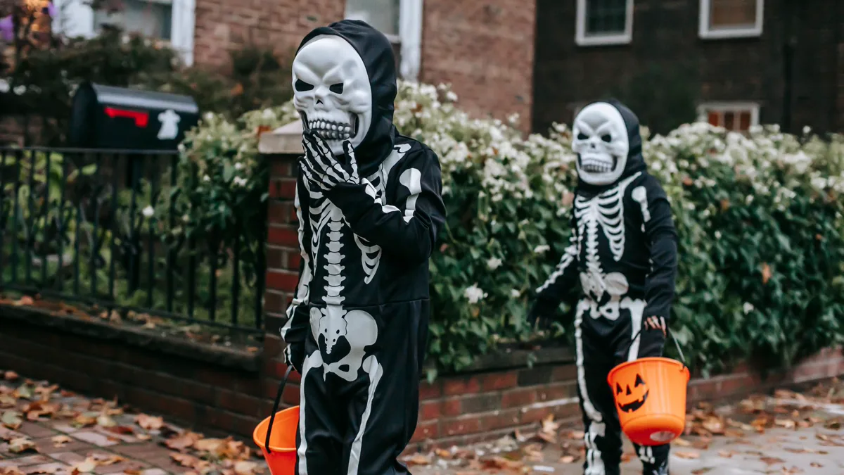 Two kids in matching skeleton suits carrying orange pumpkin buckets on a leafy sidewalk Two children in matching black skeleton costumes and masks carrying orange pumpkin buckets along a brick sidewalk covered in autumn leaves