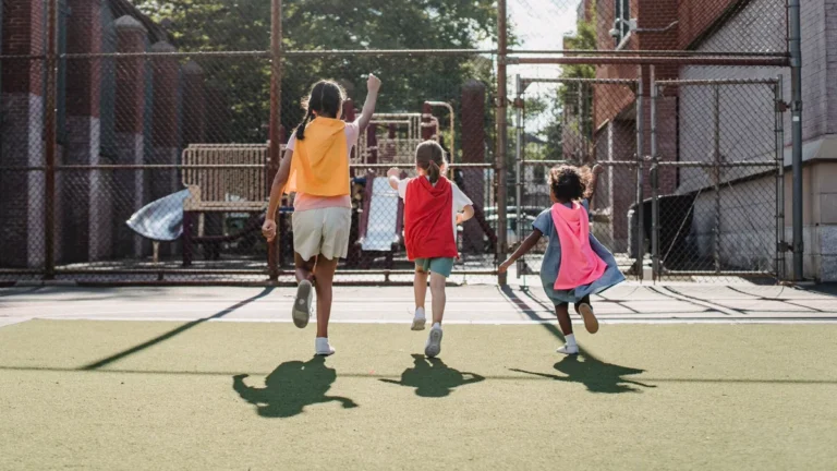 How to make a cape kids can play in: three children run across a playground wearing short, simple fabric capes