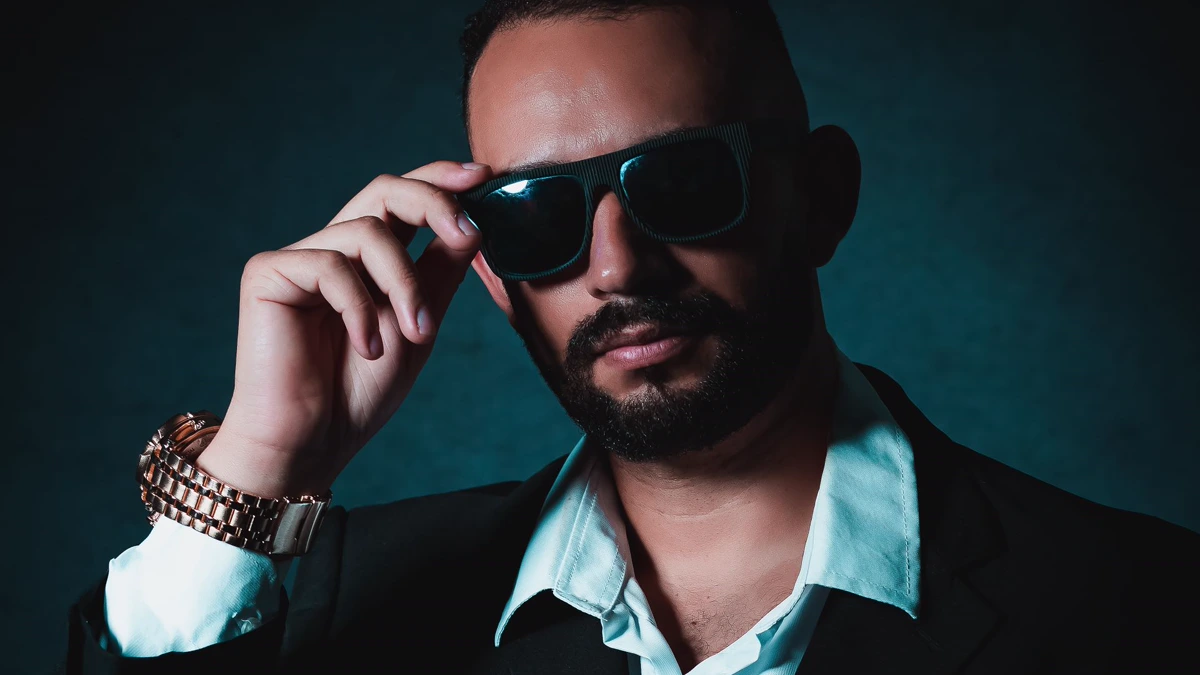 Bearded man in a dark suit jacket and open white shirt touching a pair of dark sunglasses against a teal backdrop