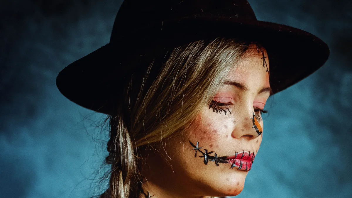 Close up of scarecrow makeup with a stitched black mouth line, drawn lower lashes, freckles and pink cheeks under a black hat