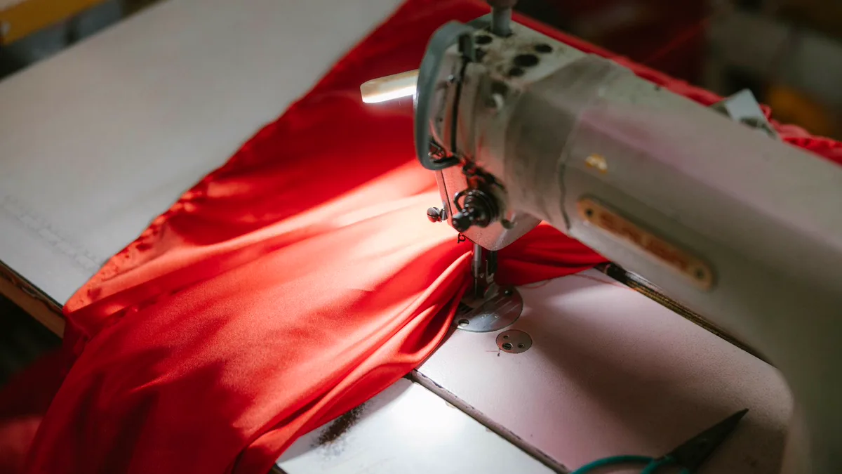 Sewing machine stitching a length of bright red satin-like fabric under a work light Sewing machine needle stitching bright red lightweight fabric, the kind of satin used for a sewn DIY cape with a narrow hem