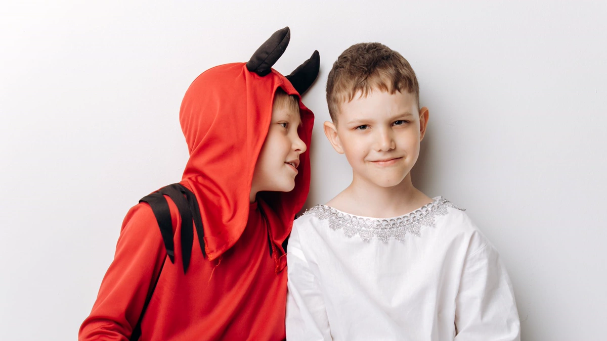 Two boys in angel and devil sibling Halloween costumes, one in a red hooded devil outfit with horns, one in a white tunic