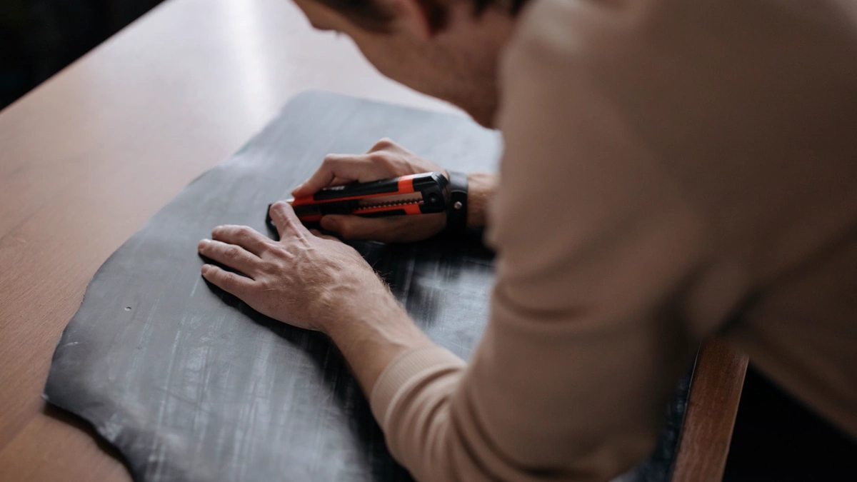 Hands holding a dark sheet flat and cutting it with an orange snap blade utility knife, the same grip used to cut cosplay armor pieces