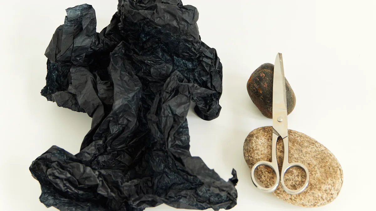 Crumpled black paper, a smooth stone and steel scissors laid out on a white table Rock, paper and scissors laid side by side, the three shapes behind one of the most recognizable trio costume ideas