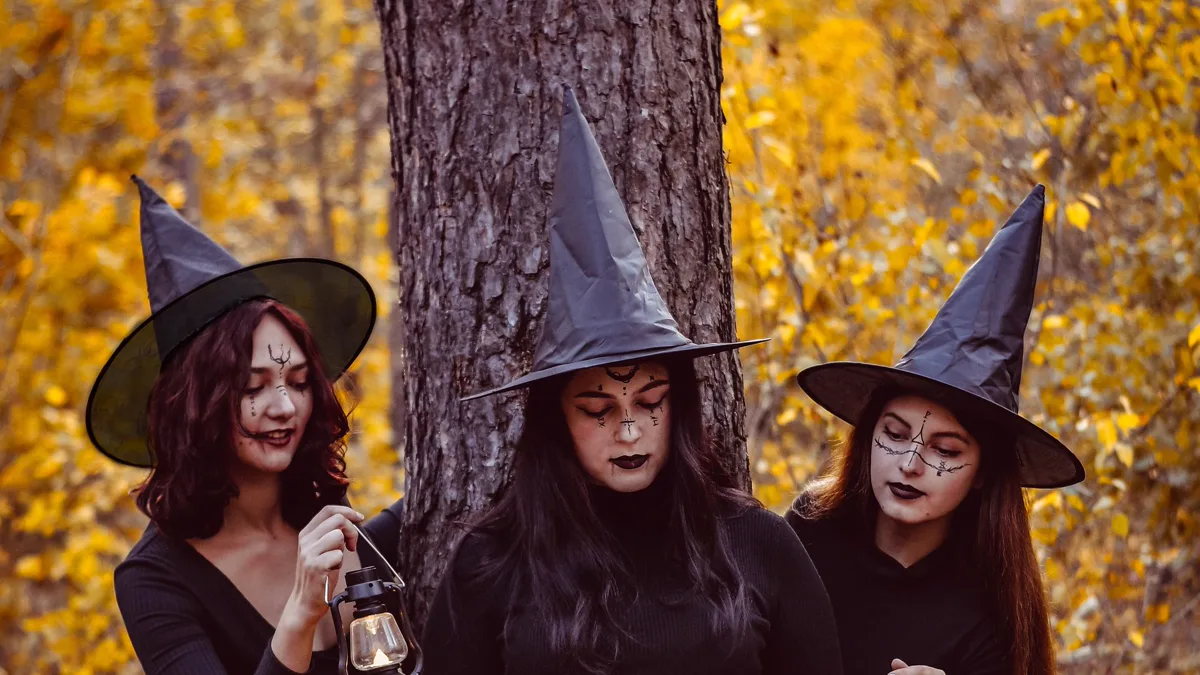 Three friends in matching black witch hats and dark outfits in an autumn forest Three women in trio Halloween costumes with matching pointed witch hats, black outfits and face paint, one holding a lantern